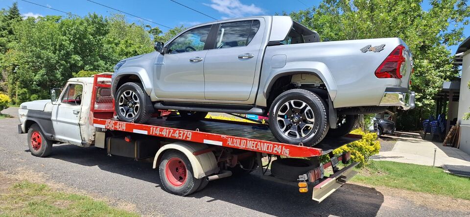 Grúa m-24 transportando una camioneta Toyota Hilux 4x4 en plataforma. Auxilio mecánico 24 horas en Argentina — teléfonos visibles en el camión.