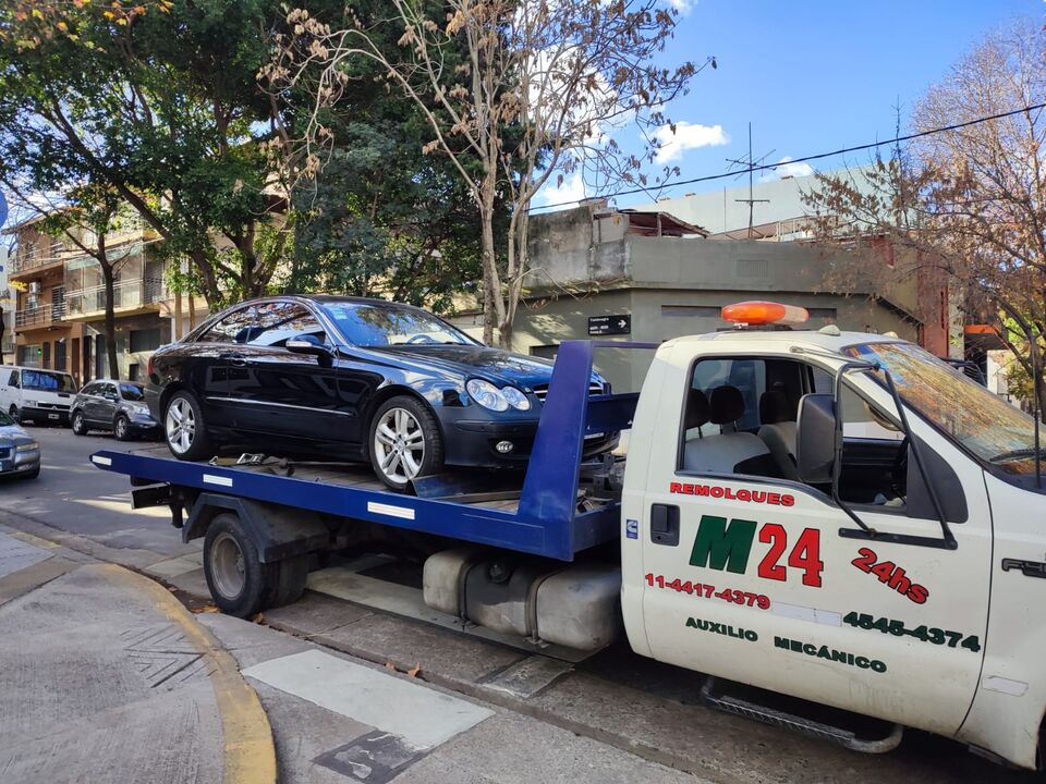 Camión remolque m-24 con logo M24 en la puerta, transportando un Mercedes-Benz CLK en calle urbana. Remolques y auxilio mecánico 24 hs.